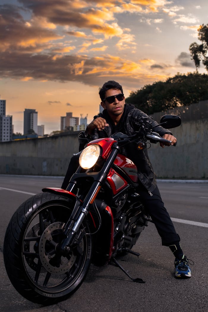 A man in sunglasses poses on a motorcycle in an urban street at sunset.