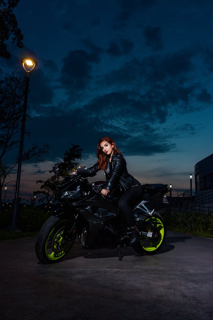 A woman in a leather jacket posing on a motorcycle at twilight, exuding confidence and style.