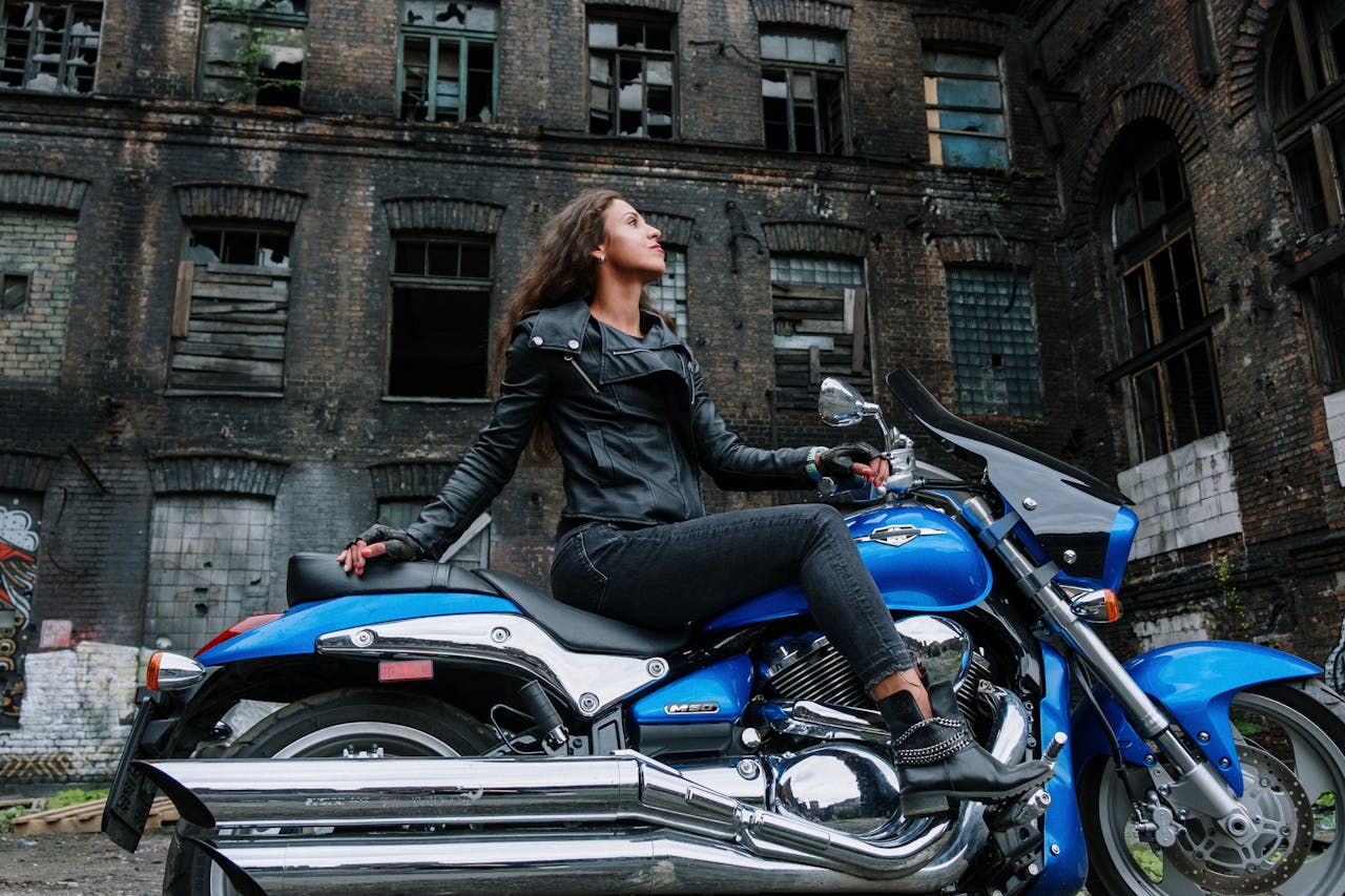 A woman in a black leather jacket sits confidently on a blue motorcycle in front of an abandoned building.
